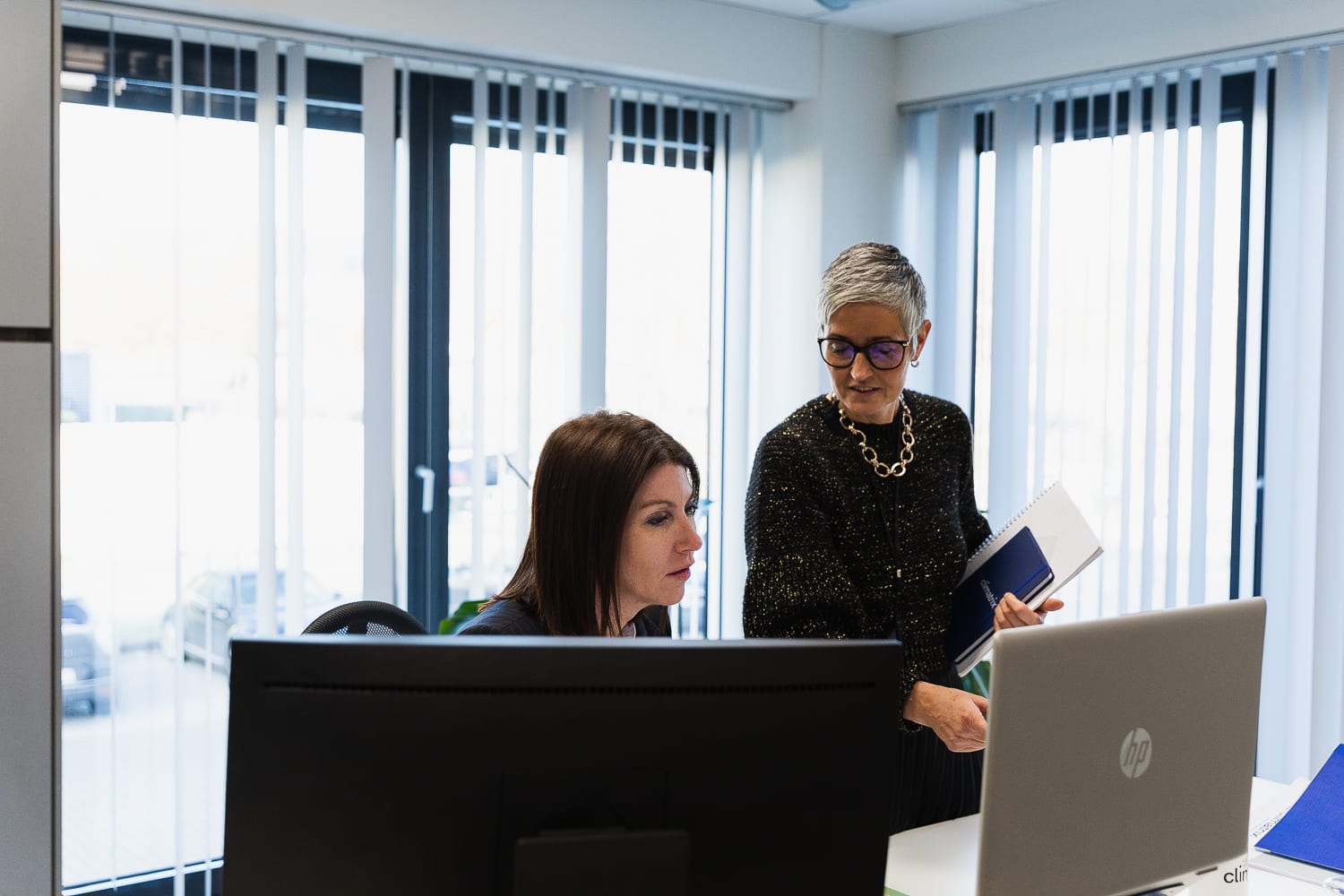 Zaakvoerder Kim bekijkt samen met een vrouwelijke collega enkele zaken op haar laptop. Ze zit achter haar bureau, haar collega staat naast haar en heeft een schrift vast.